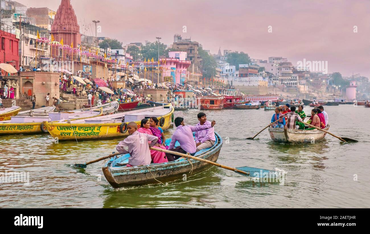 Indian turisti essendo remato lungo il fiume Gange, passando per il lungomare ghats riempito di pellegrini Indù che si bagnano nel fiume santo. Foto Stock
