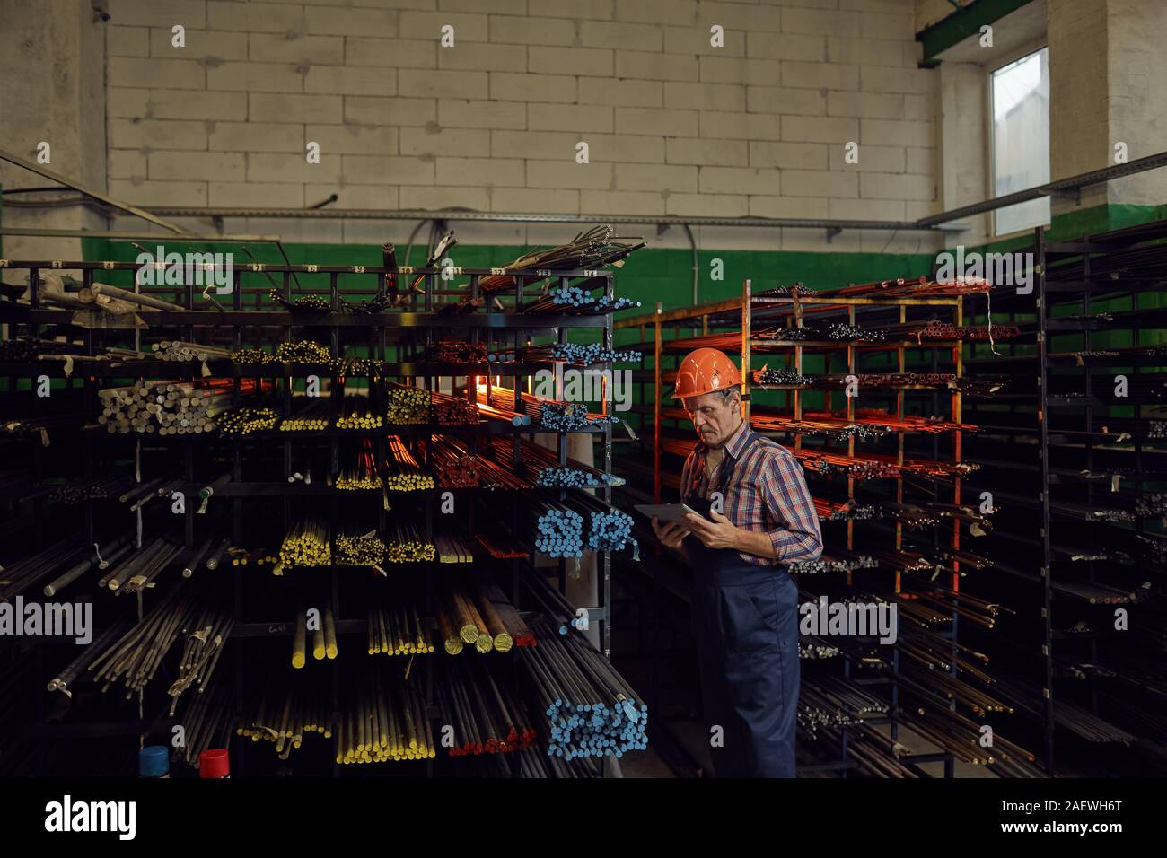 Grave senior uomo nel casco di lavoro controllare l'ordine online su tablet PC mentre sta in piedi vicino a tubi di metallo in magazzino Foto Stock