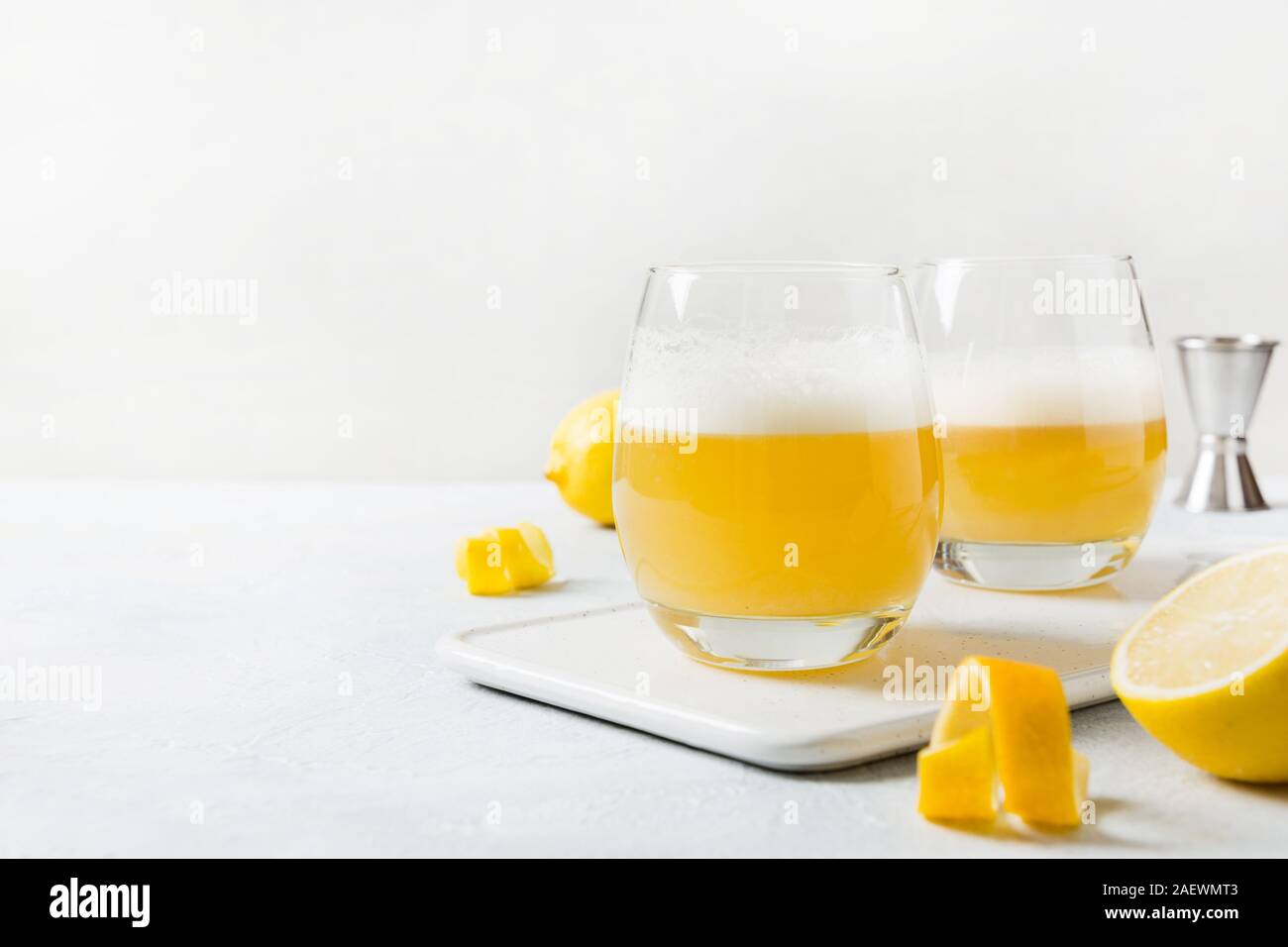 Due whiskey sour cocktail con gli ingredienti, il succo di limone e lo sciroppo di zucchero e albume in vetro sul tavolo bianco. Orientamento orizzontale. Foto Stock