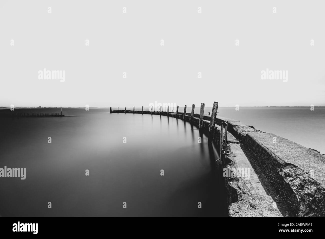 Calcestruzzo stagionato frangionde con un foglio arrugginito palo angolata e travi in legno riflessa nello specchio di acqua liscia superficie su un windless giornata con un bl Foto Stock
