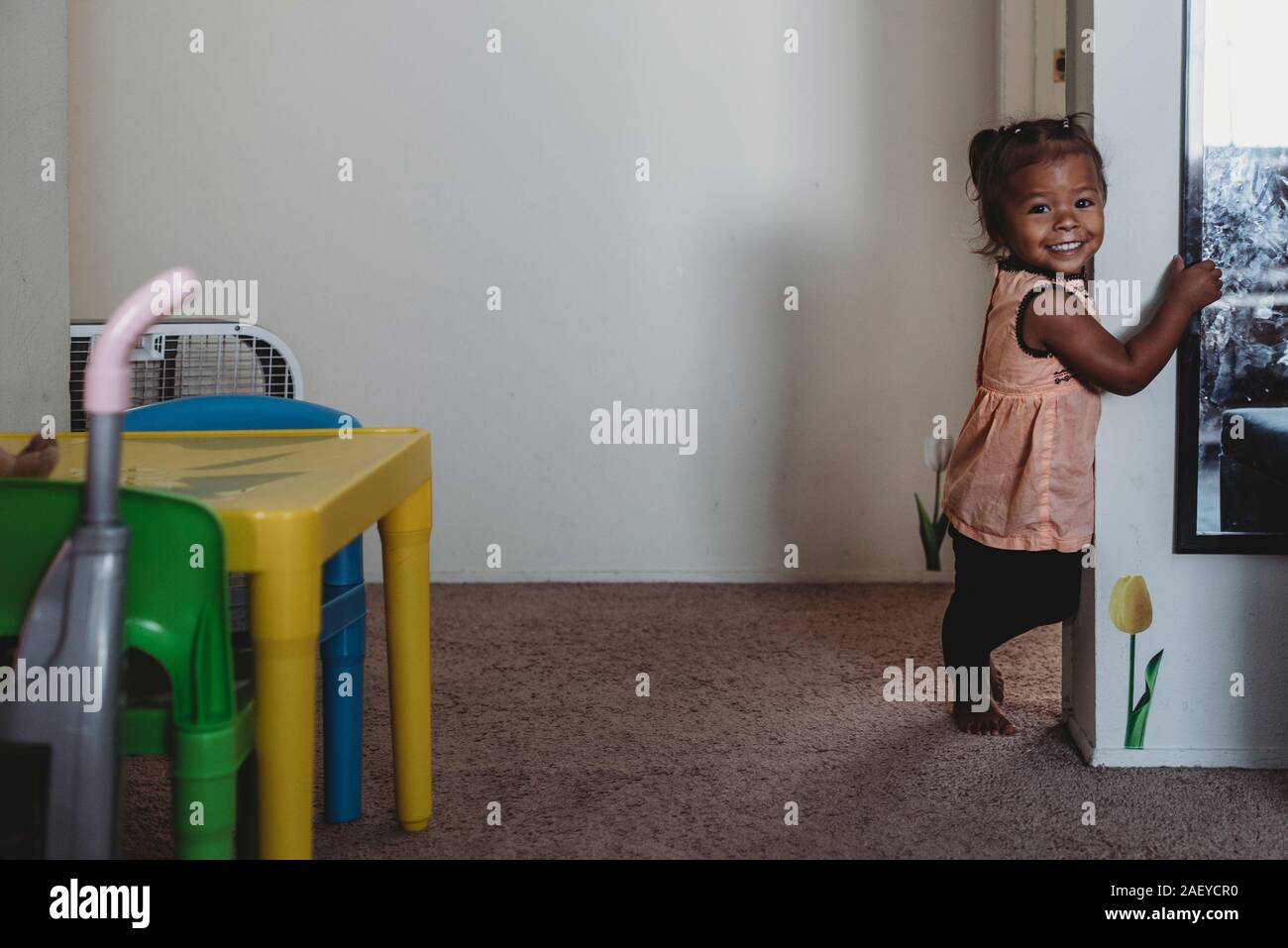 Sorridente multirazziale 2 yr old girl in piedi su un tappeto accanto alla parete Foto Stock