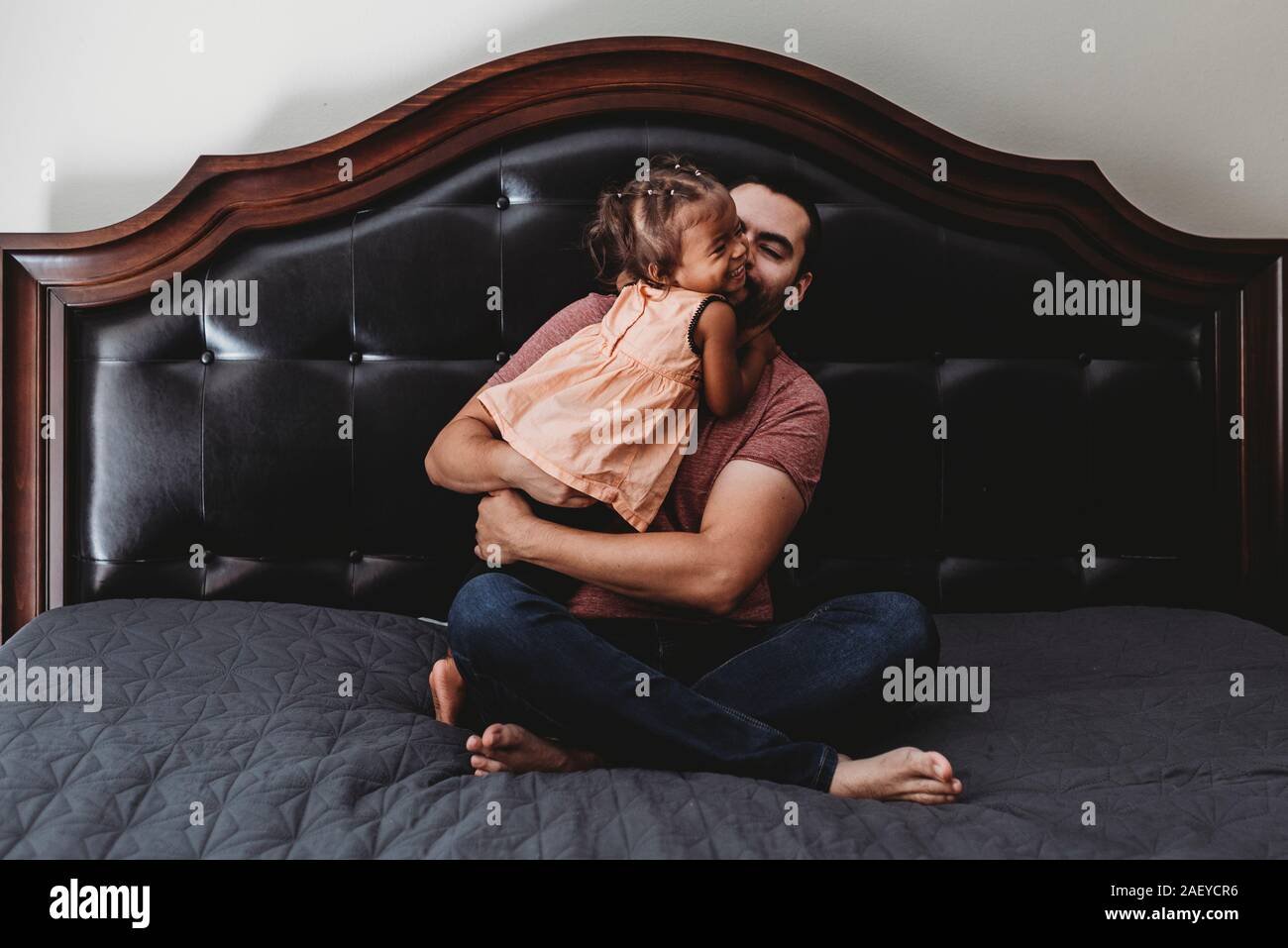 A piedi nudi papà seduta sul letto di ridere con 2 anni figlia biracial Foto Stock