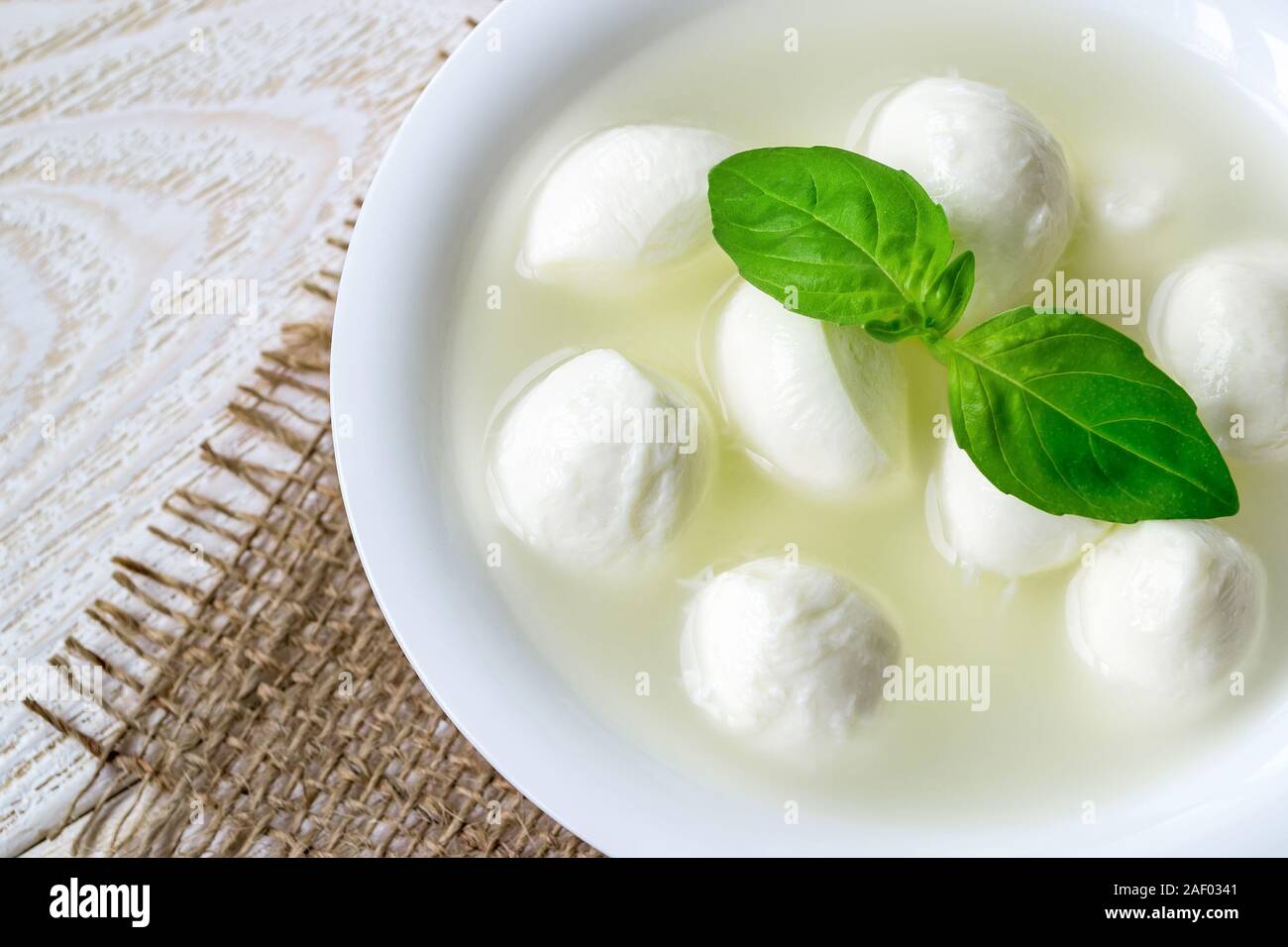 Close-up di ciotola con mini mozzarella di palline in una soluzione salina e basilico su un bianco tavola in legno rustico. Healhy cucina mediterranea. Foto Stock
