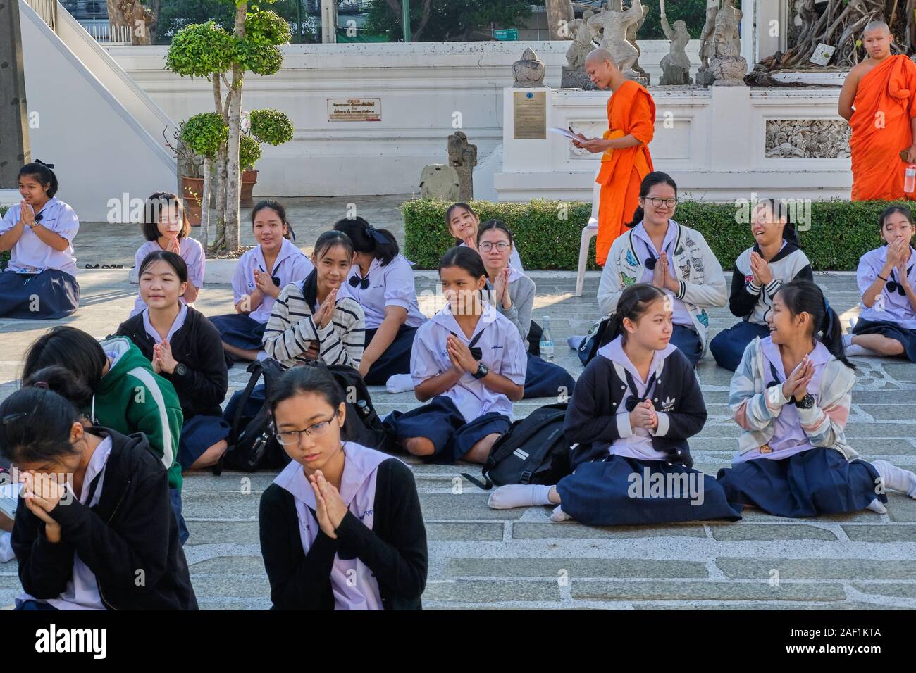 Scuole di Thai ragazze nella motivazione di Wat Suthat di Bangkok, Tailandia, essendo indicato da due monaci buddisti in preghiera buddista Foto Stock