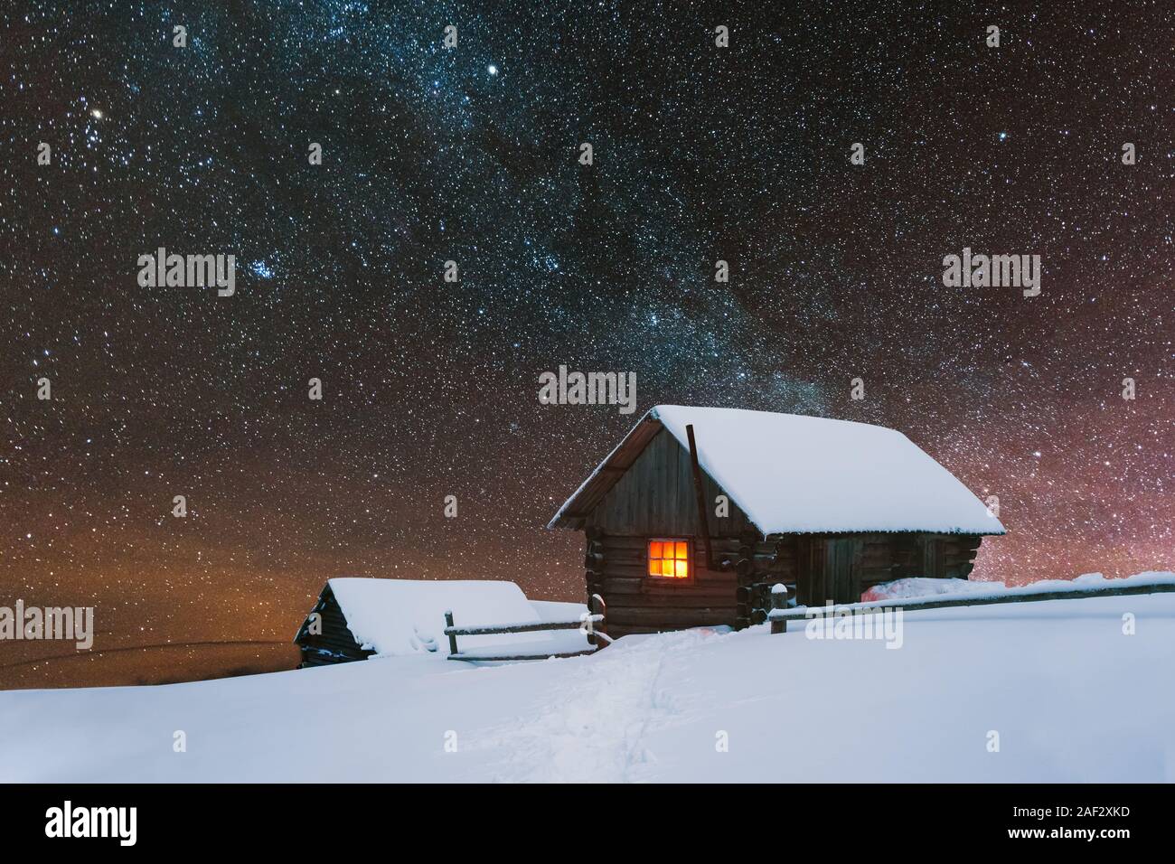 Drammatica scena invernale con casa innevato con una luce calda nella finestra . Fantastico paesaggio notturno incandescente dalla via lattea. Inverno in alta montagna Foto Stock