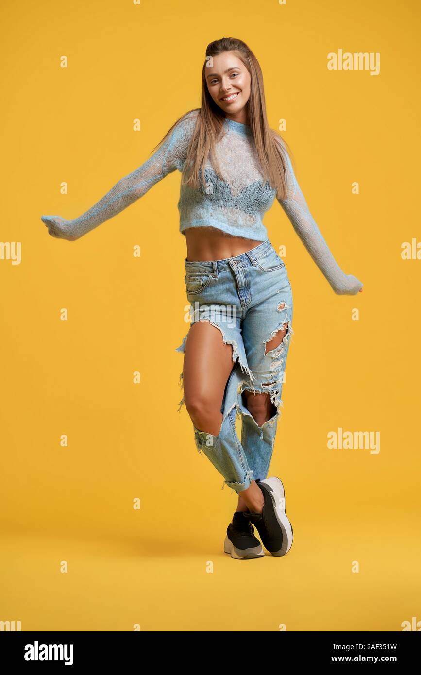 Vista frontale della elegante attraente ragazza in jeans strappati e  maglione blu. Bella bruna giovane donna con i capelli lunghi e il sorriso  sincero che posano su sfondo giallo. Concetto di divertimento,