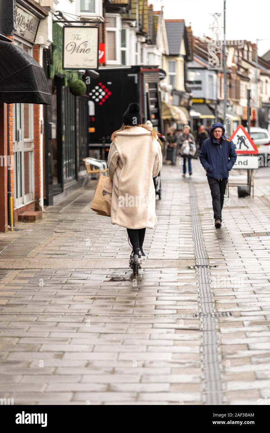 Equitazione femmina uno scooter elettrico sul marciapiede in Leigh on Sea, Essex, Regno Unito vicino a una passeggiata pedonale. Wet Rainy day. Nessun casco. Mezzi personali di trasporto Foto Stock