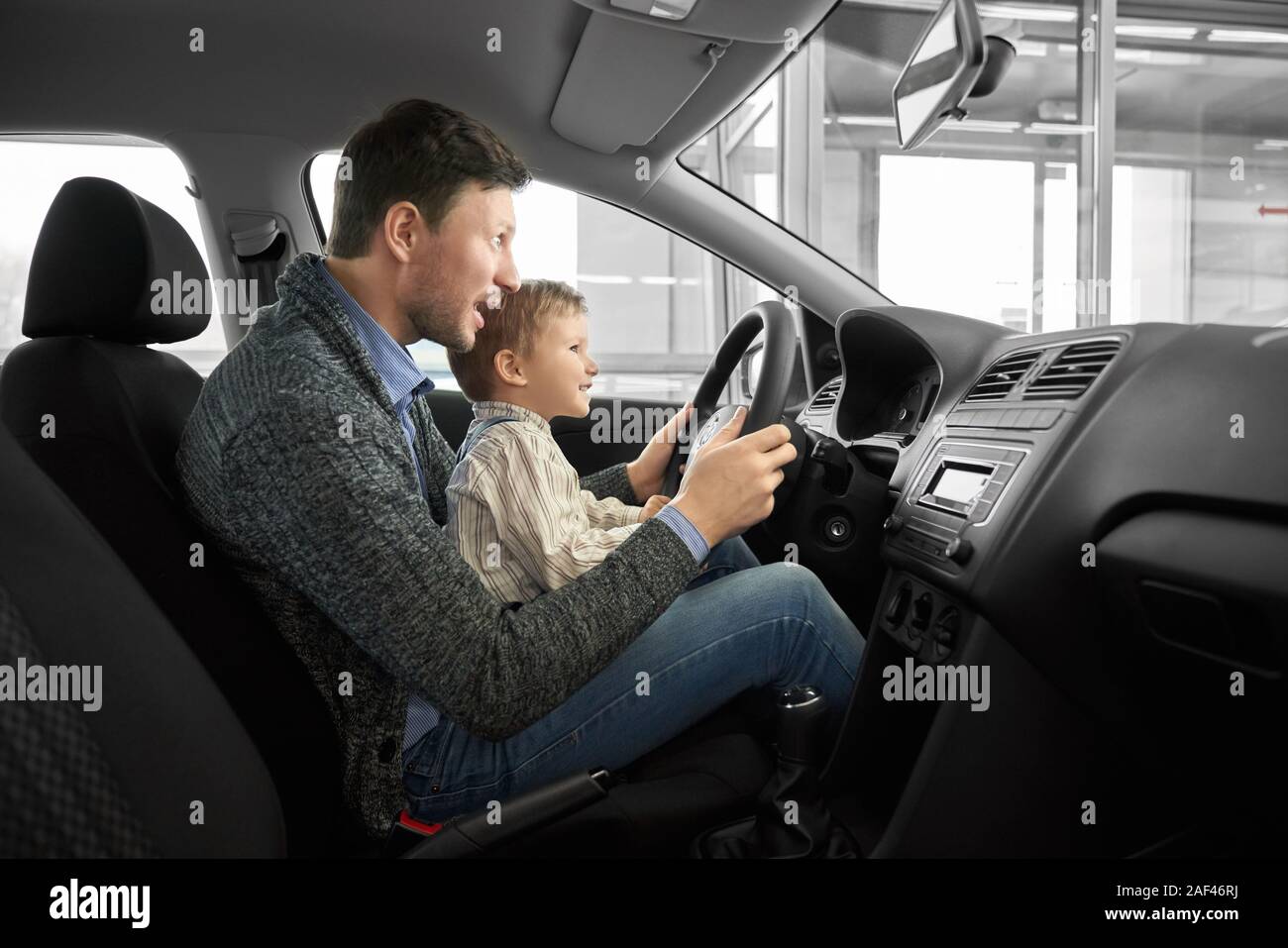 Vista dal lato di divertenti papà e figlio seduto sul sedile del driver nella nuova auto,mantenendo le mani sul volante e guardando in avanti. Client Test di macchine e la scelta di automobile. Concetto di auto salon. Foto Stock