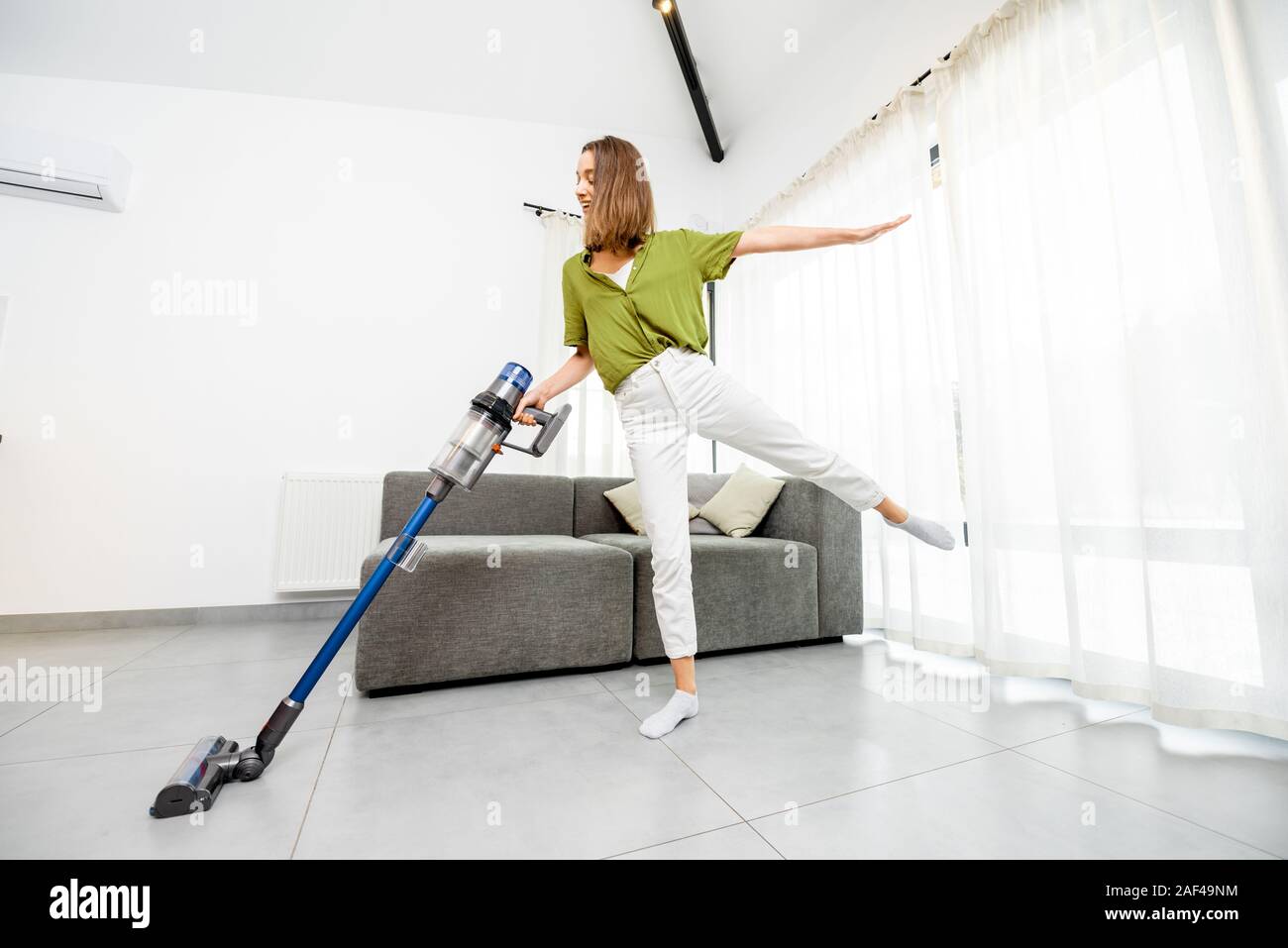 Giovane donna godendo i lavori domestici, pulizia pavimento cordless con aspirapolvere in bianco moderno salotto. Concetto di facilità di pulizia con un wireless aspirapolvere Foto Stock