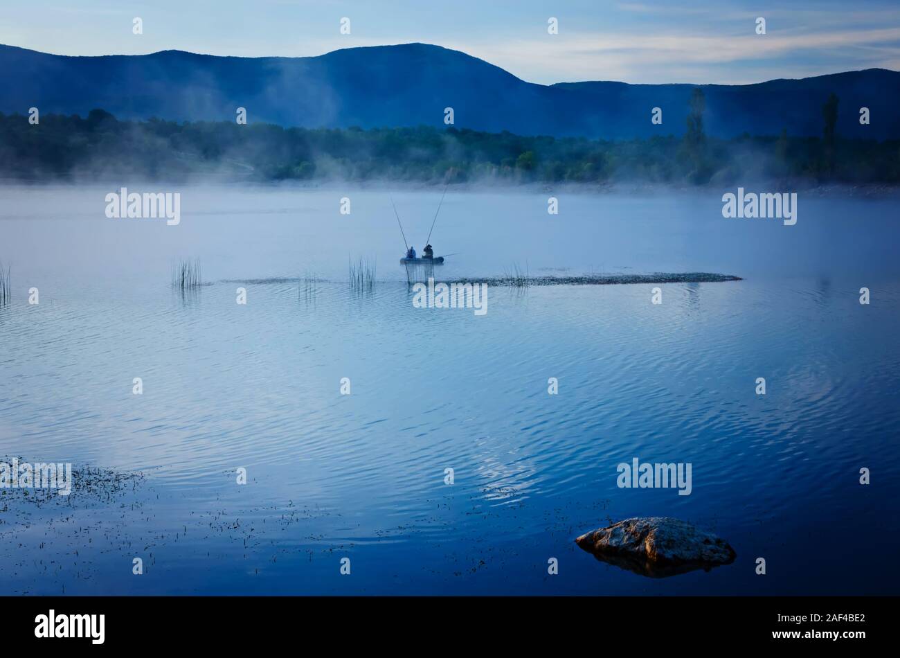 Nebbia mattutina sul lago. Sagome di due uomini con attività di pesca in una barca. Il paesaggio nel quartiere alla moda di toni del blu. Colore dell'anno 2020, classic blu. Foto Stock