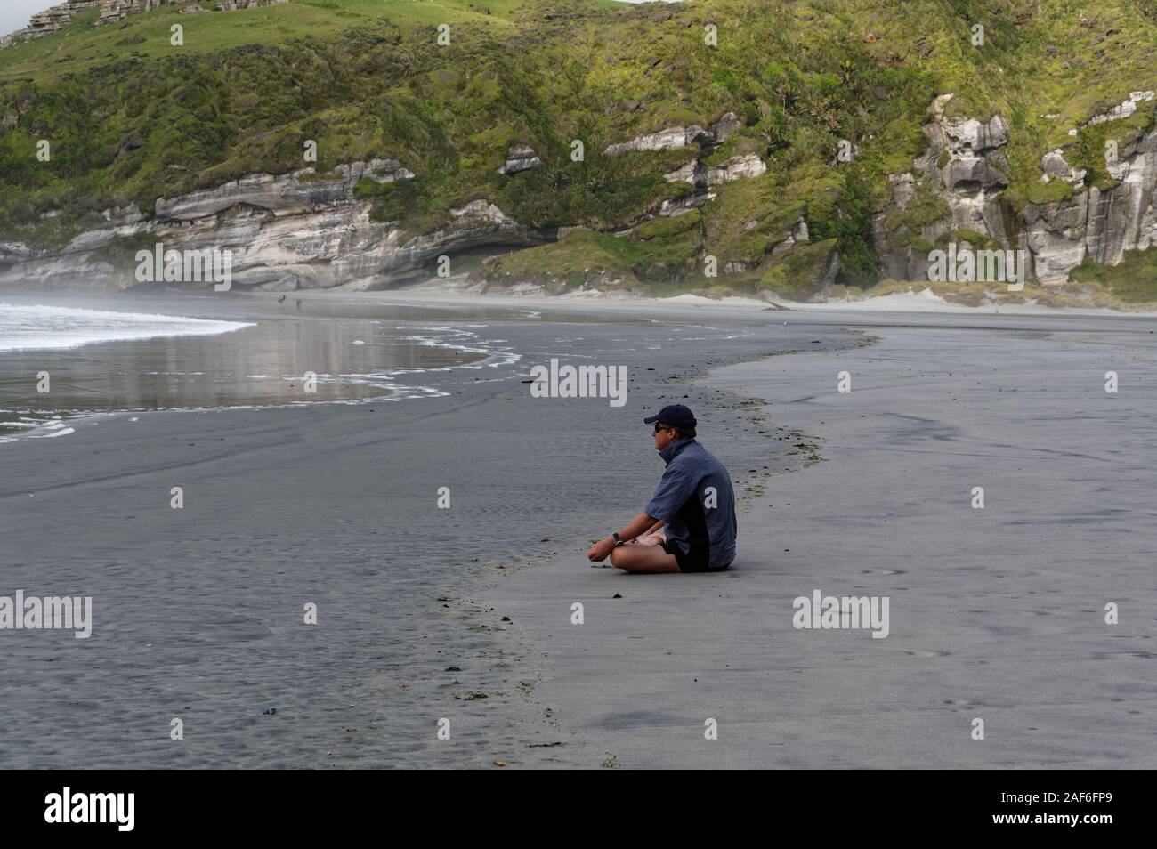 Un uomo è seduto zampe trasversale sulla spiaggia come le onde giro verso di lui. Foto Stock
