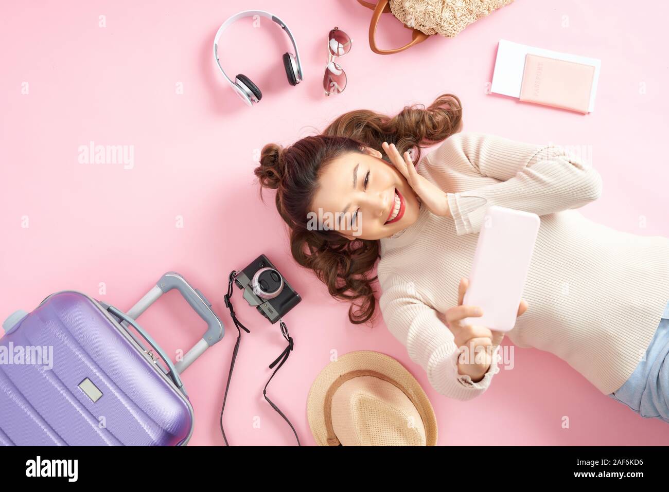 Felice ragazza asiatica usando il telefono per scattare foto e giacente sul piano di colore rosa con bagaglio da viaggio. Vista superiore Foto Stock