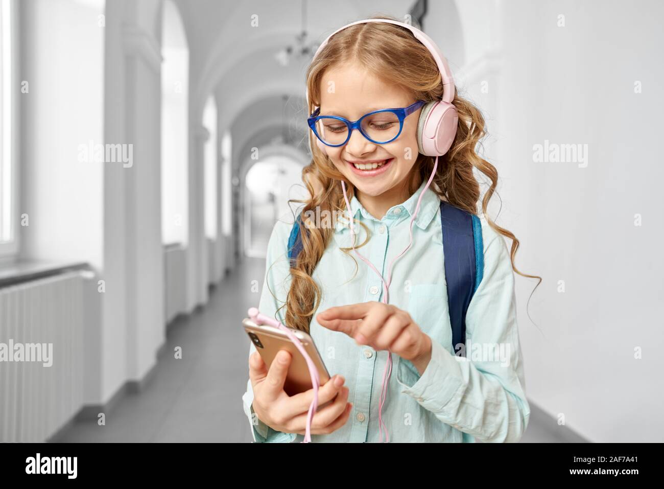 Bella piccola scuola ragazza con i riccioli biondi indossando occhiali e camicia blu utilizza lo smartphone e sorridente. Bella ragazza sul corridoio, chiamata in attesa Foto Stock