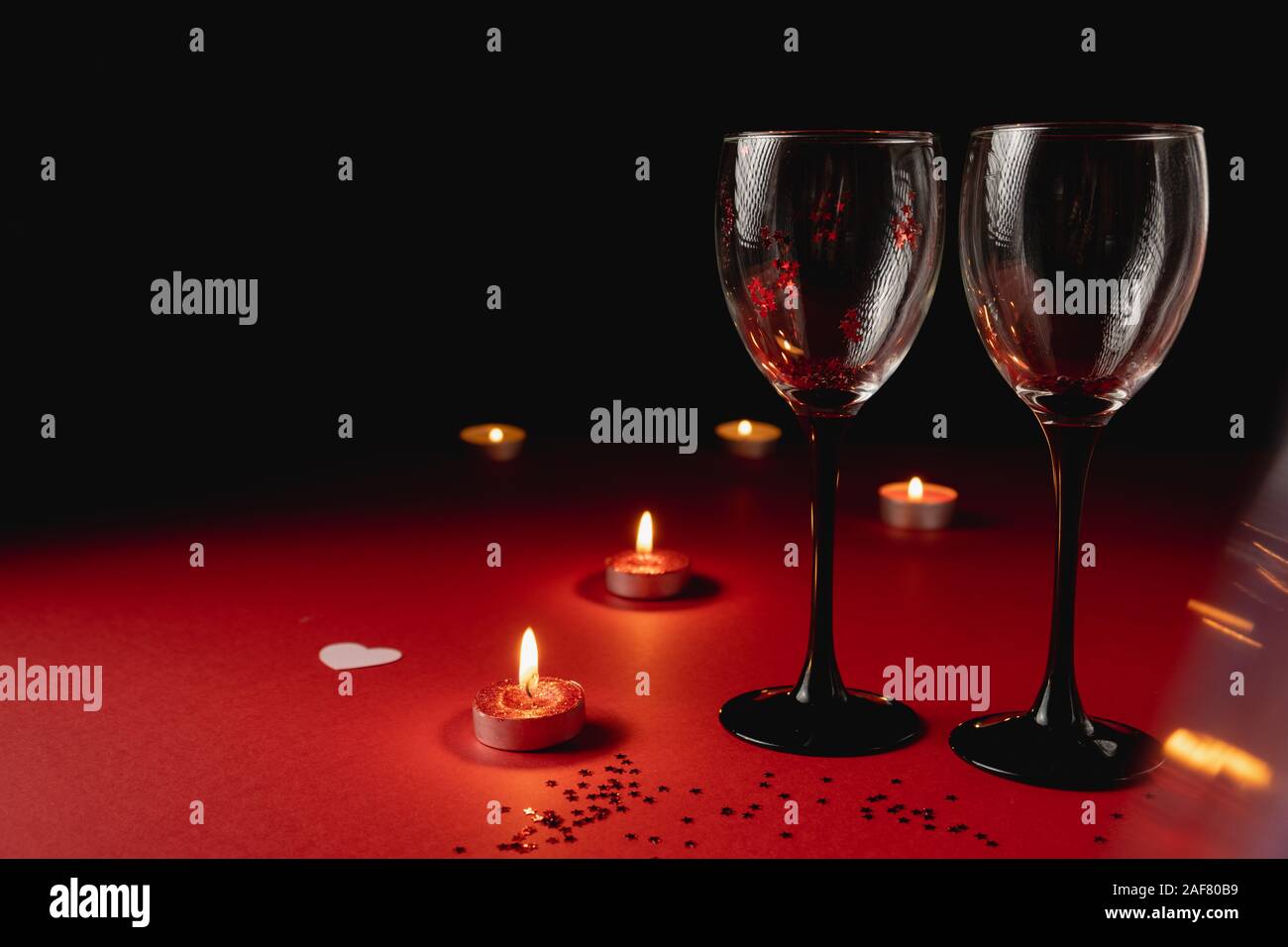 Sfondo di vacanza. Il tintinnio di due bicchieri di vino, candela, coriandoli sul rosso e sfondo nero. Stile minimal e il giorno di San Valentino del concetto. Laici piana, copia Foto Stock