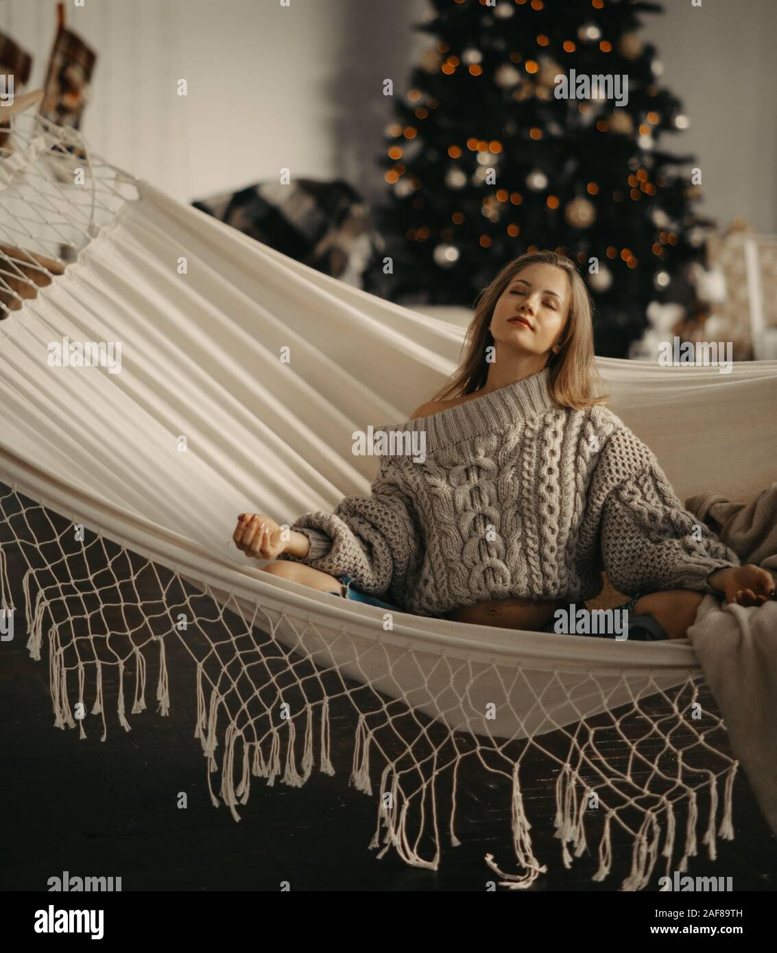 Giovane donna incinta in pullover si siede e medita nel yoga posa in amaca sullo sfondo di albero di Natale . Foto Stock