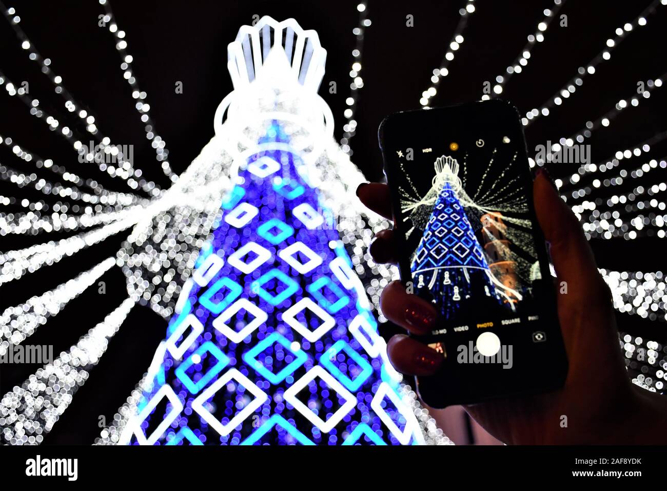 Albero di Natale decorato di bianco e blu con luci di Natale 2019 e Capodanno 2020, mercato e celebrazioni a Vilnius, ragazza canto e lo smartphone Foto Stock