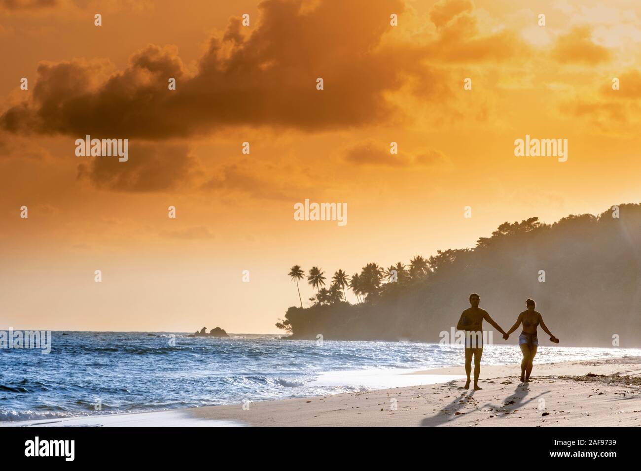 Una giovane coppia tenendo le mani e correndo insieme lungo una spiaggia tropicale contro un tramonto dorato Foto Stock