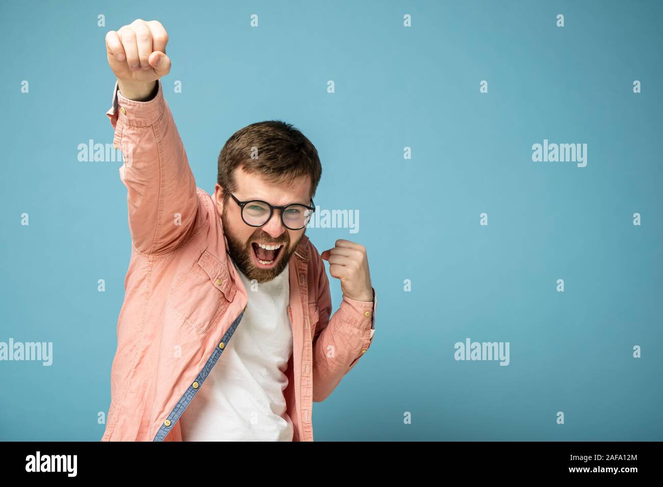 Gioioso e sorridente uomo barbuto alza la mano, sensazione di vittoria e successo. Isolato su uno sfondo blu. Foto Stock