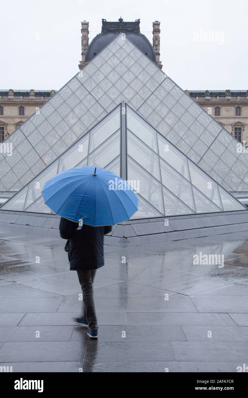 Un uomo con un ombrello blu al di fuori del Louvre e la famosa piramide di vetro ingresso in Parigi Foto Stock