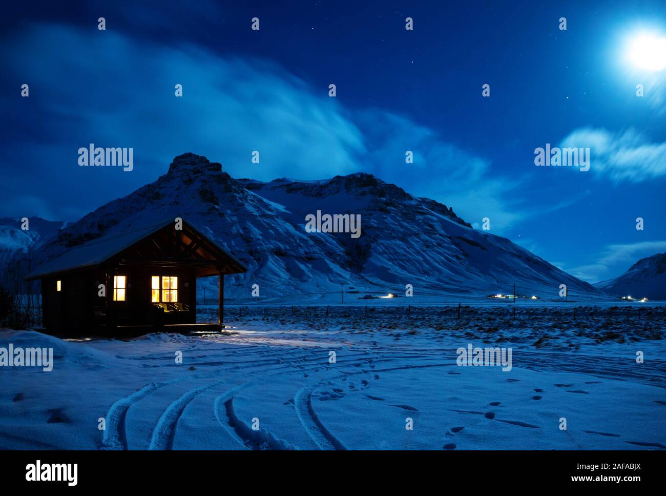 Paesaggio con una casa in legno con la luce dalla finestra in una notte d'inverno. Vista panoramica del chiaro di luna sulla neve con le montagne sullo sfondo. Così Foto Stock