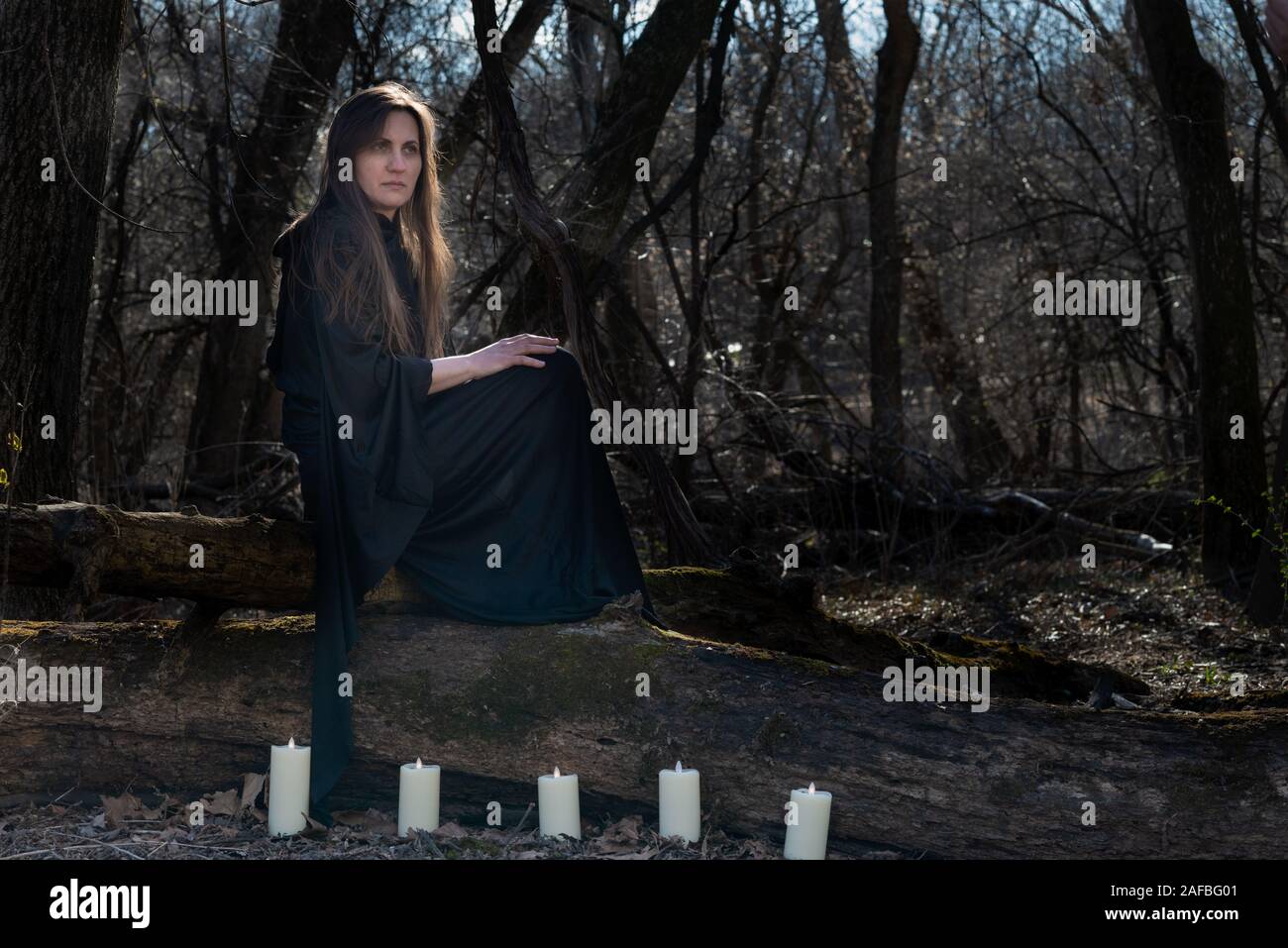 Donna con scuri capelli lunghi in black robes circondato da white candele accese seduto su un tronco di albero nella foresta. Torna al concetto di natura. Stregoneria Foto Stock