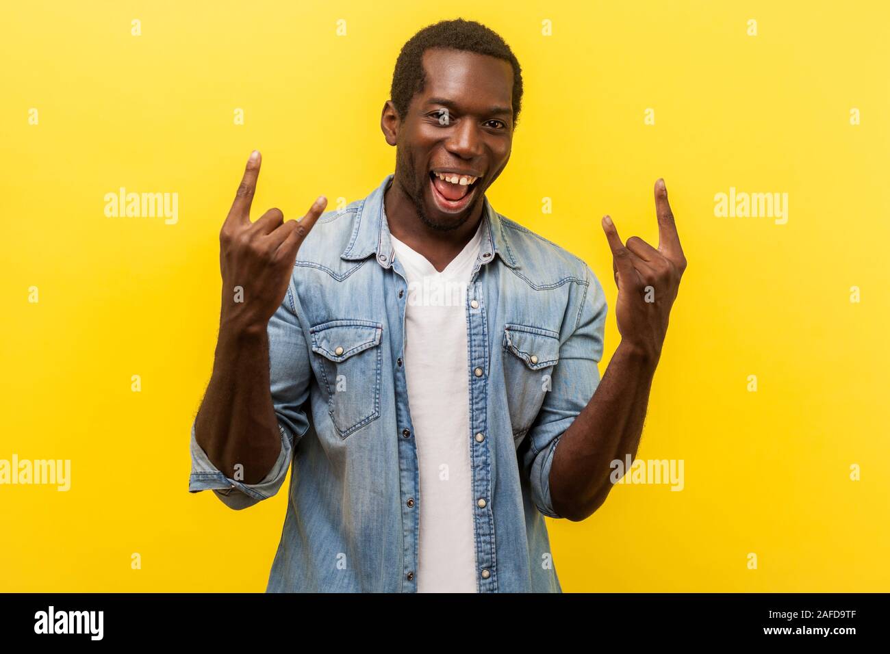 Ritratto di entusiasti crazy allegro uomo in denim Camicia casual con arrotolato a maniche che mostra rock and roll gesti con le mani fino, sorridente alla fotocamera. in Foto Stock