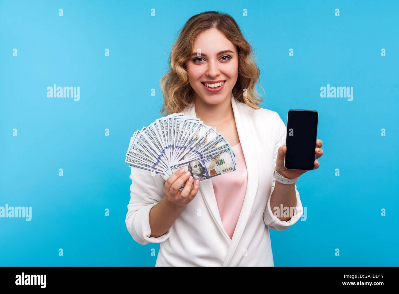 Pagamento on line, transazioni di denaro. Ritratto di donna positiva con capelli ondulati in camicia bianca mostra di dollari e il telefono cellulare, vantando earni internet Foto Stock