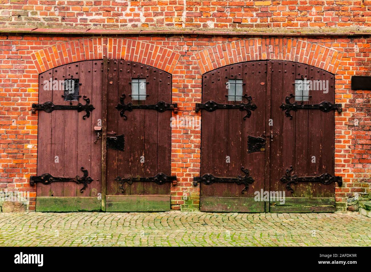 Chiudere l immagine di una coppia di porte in legno di uno storico edificio di mattoni rossi, con supporti in metallo e serrature con moss sulla parete e sul terreno. Foto Stock