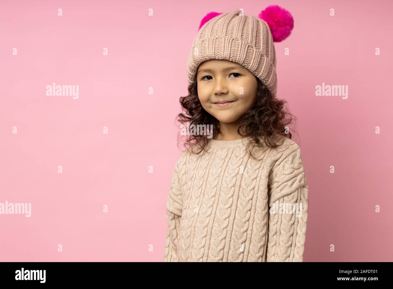 Ritratto orizzontale di amichevole bambina con capelli ricci, indossa un  maglione beige, Cappello invernale con crimson peloso pom pom, sorridente  guardando la telecamera Foto stock - Alamy