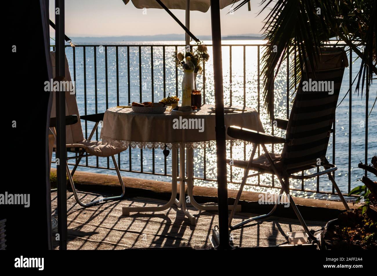 Servite la colazione tavola per due con due trasferte sedie e ombrellone dietro le ringhiere metalliche sulla spiaggia del lago di Garda a fine sunrise. Lombardia, Ita Foto Stock