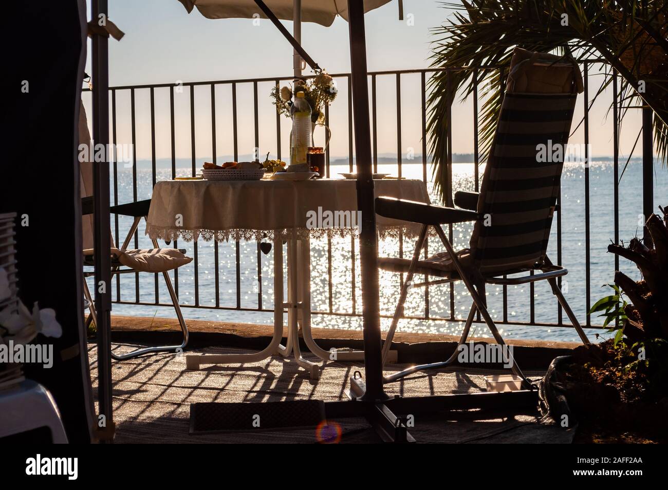 Servite la colazione tavola per due con due trasferte sedie e ombrellone dietro le ringhiere metalliche sulla spiaggia del lago di Garda a fine sunrise. Lombardia, Ita Foto Stock