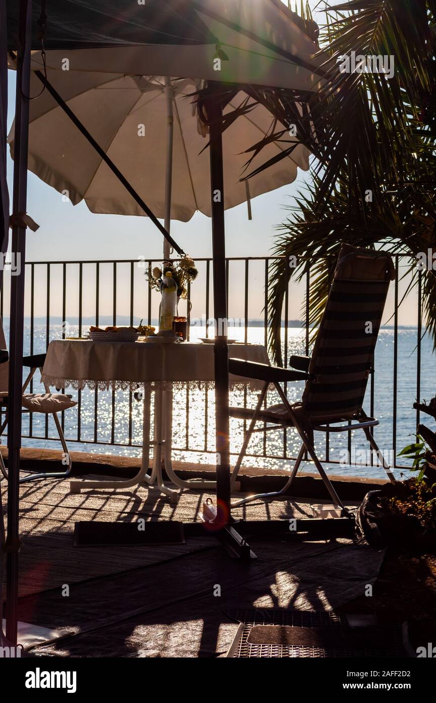 Servite la colazione tavola per due con due trasferte sedie e ombrellone dietro le ringhiere metalliche sulla spiaggia del lago di Garda a fine sunrise. Lombardia, Ita Foto Stock