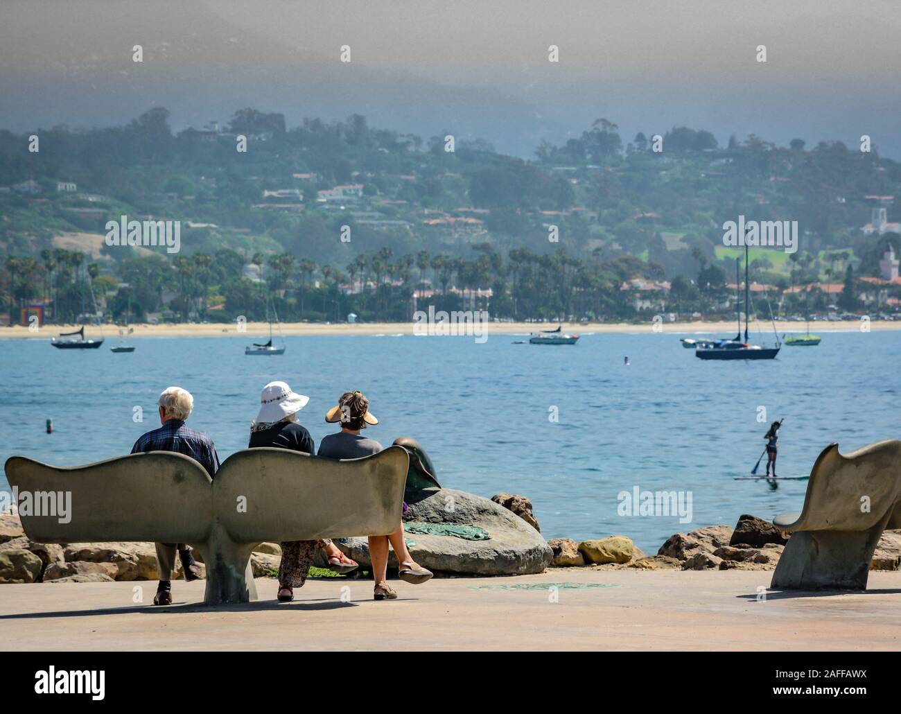 Persone sedersi sulla coda di balena panca si affaccia la chiesa di Santa Barbara del porto con SUPB, barche a vela e le prime colline nella nebbia, a Santa Barbara, CA, Stati Uniti d'America Foto Stock