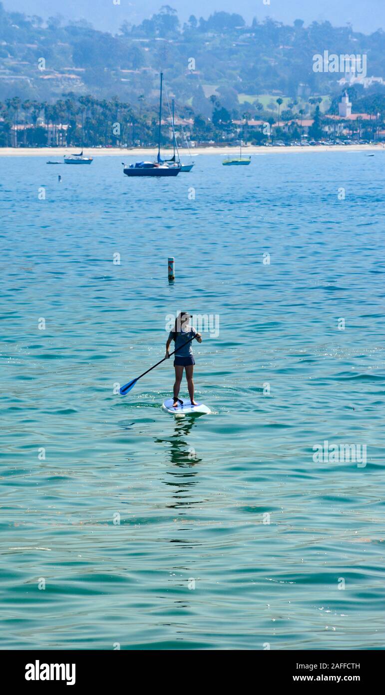 Una solitaria giovane donna pagaie il suo Stand Up Paddleboard attraverso la Santa Barbara porto vicino barche a vela e la spiaggia di Santa Barbara, CA, Foto Stock