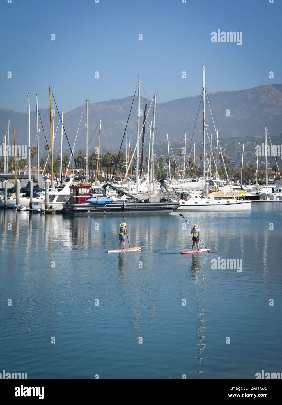 Due donne maneuverer loro stand up paddle boards attraverso un canale vicino ormeggiata barche a vela e yacht in marina nella Santa Barbara, Porto Santa Foto Stock