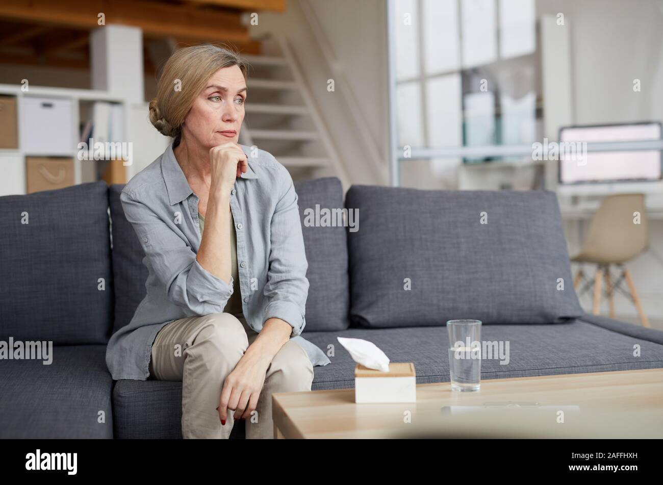 Ritratto di eleganti donna matura seduti sul divano di casa e guardando lontano pensively, triste o depressa con tessuti su tavola, spazio copia Foto Stock