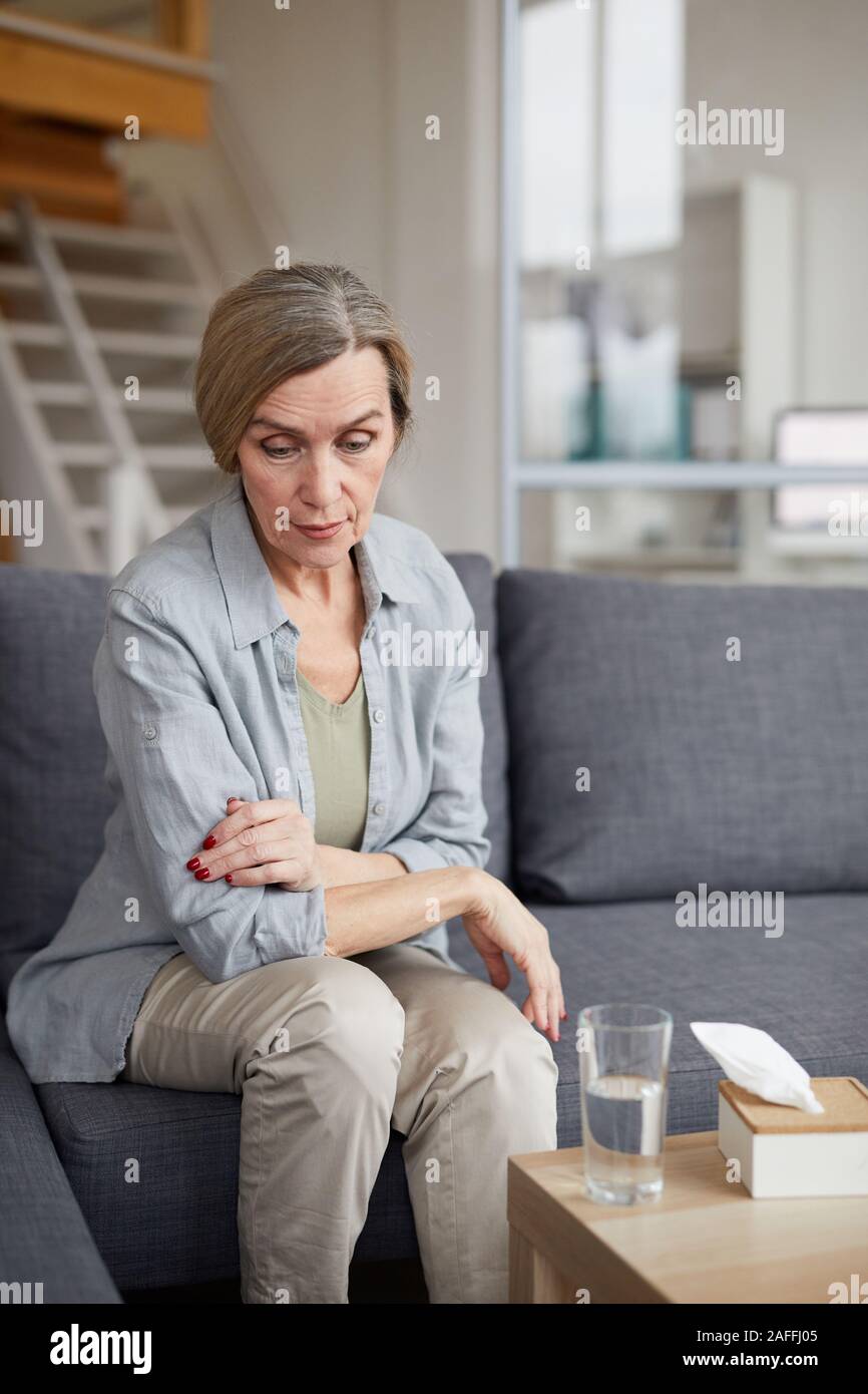 Ritratto di triste donna matura seduti sul divano di casa e guardando verso il basso con la faccia triste espressione, tessuti e acqua sul tavolo, spazio di copia Foto Stock