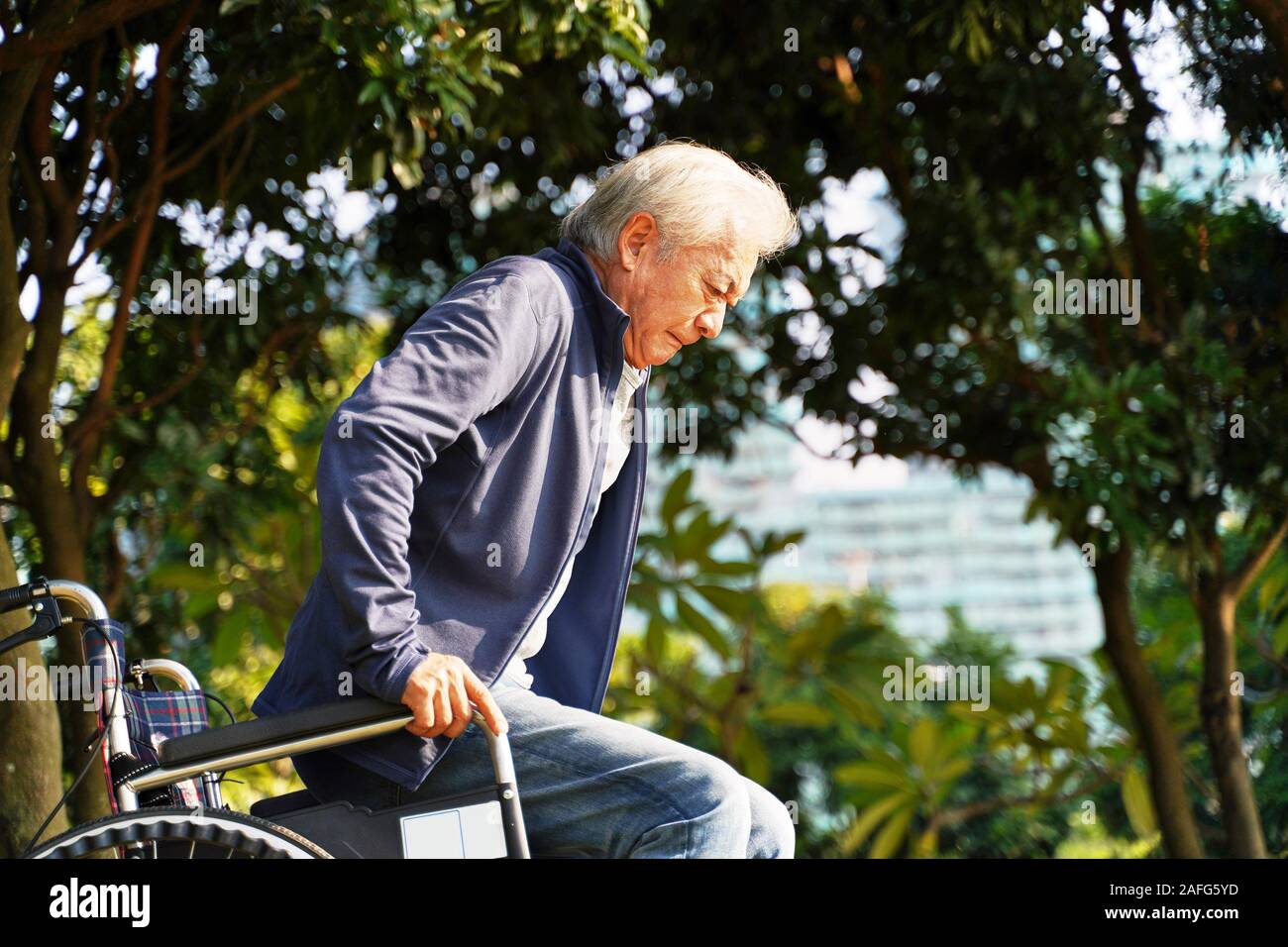 Senior uomo asiatico cercando di alzarsi dalla sedia a rotelle all'aperto Foto Stock