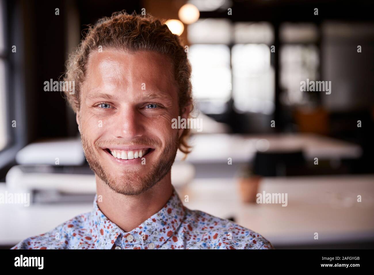 Millenaria maschio bianco creative seduta sorridente in ufficio alla fotocamera, la testa e le spalle da vicino Foto Stock