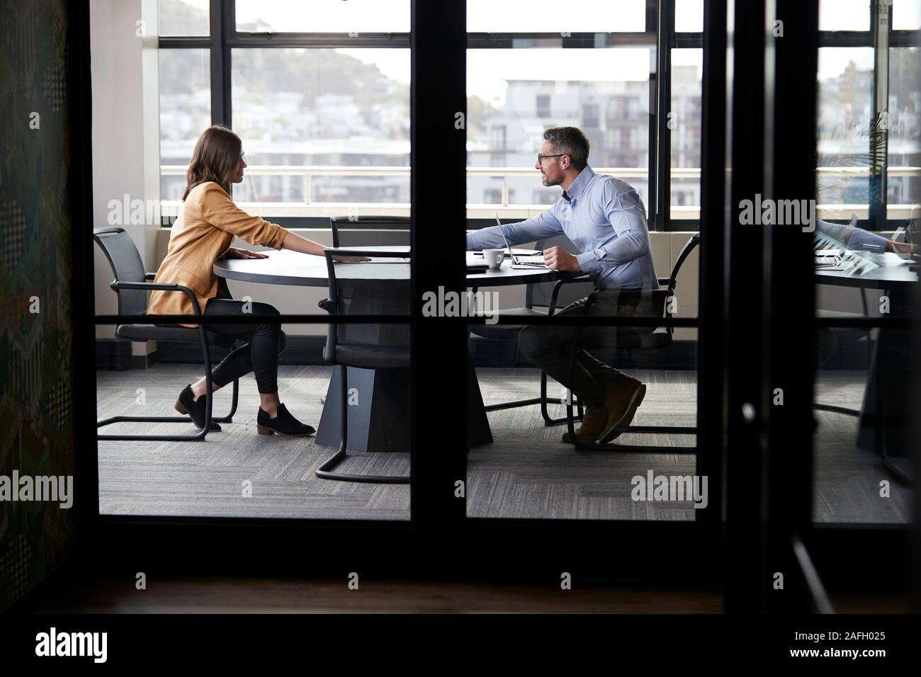 Un imprenditore e la giovane donna incontro per un colloquio di lavoro a piena lunghezza e visto attraverso la parete in vetro Foto Stock