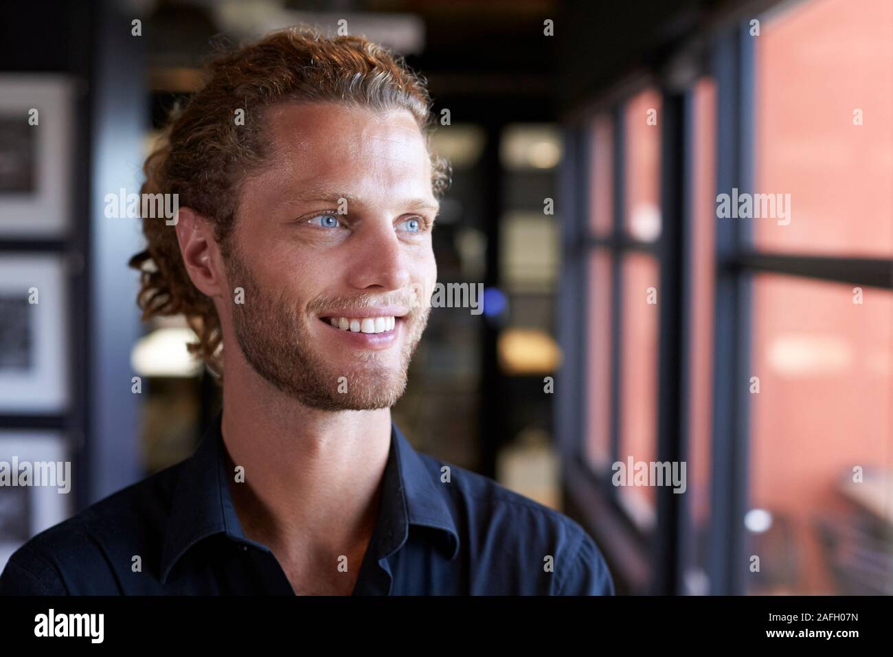 La millenaria maschio bianco creative business sorridente e guardando fuori della finestra in un ufficio, close up Foto Stock