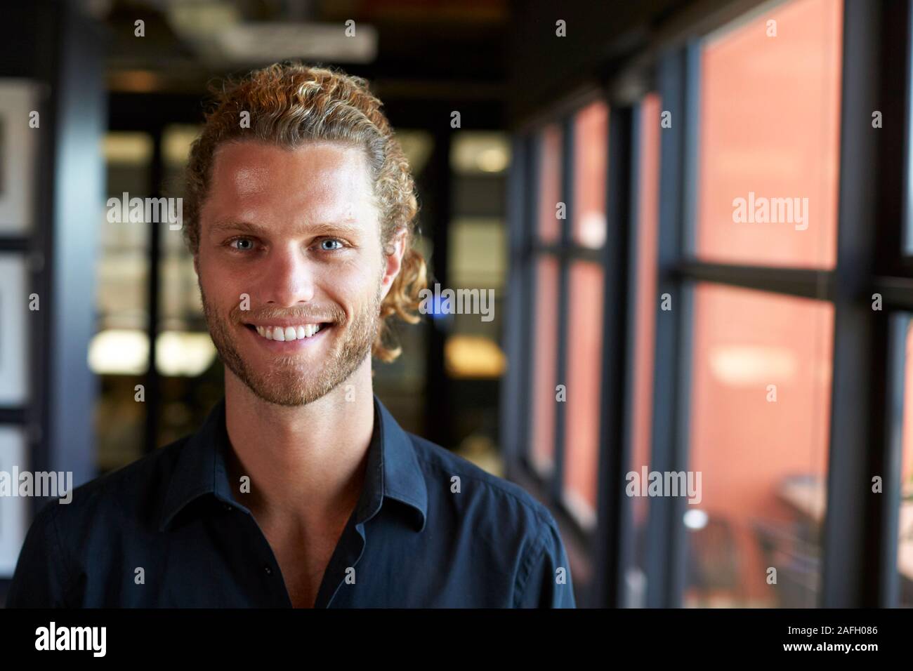 La millenaria maschio bianco creative business sorridere alla telecamera tramite la finestra in un ufficio, close up Foto Stock