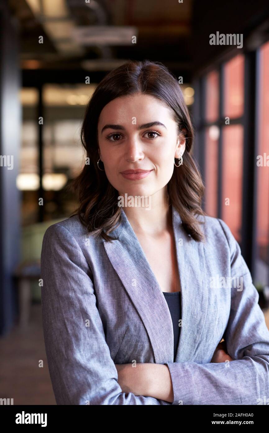 Bianco millenario imprenditrice in piedi con le braccia incrociate di sorridere alla telecamera in un ufficio, close up Foto Stock
