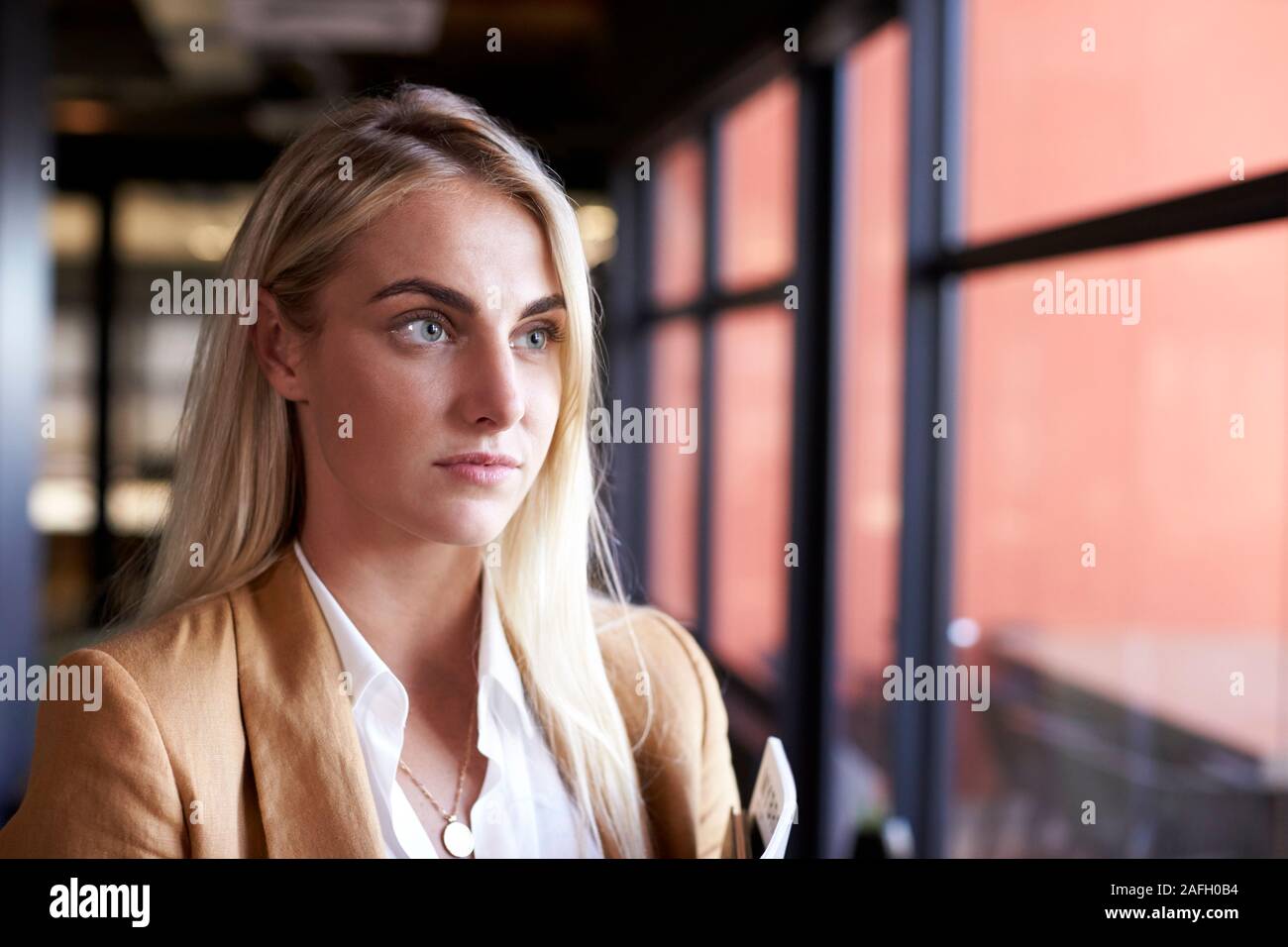 Bianco millenario bionda imprenditrice guardando fuori della finestra in un ufficio, testa e spalle Foto Stock