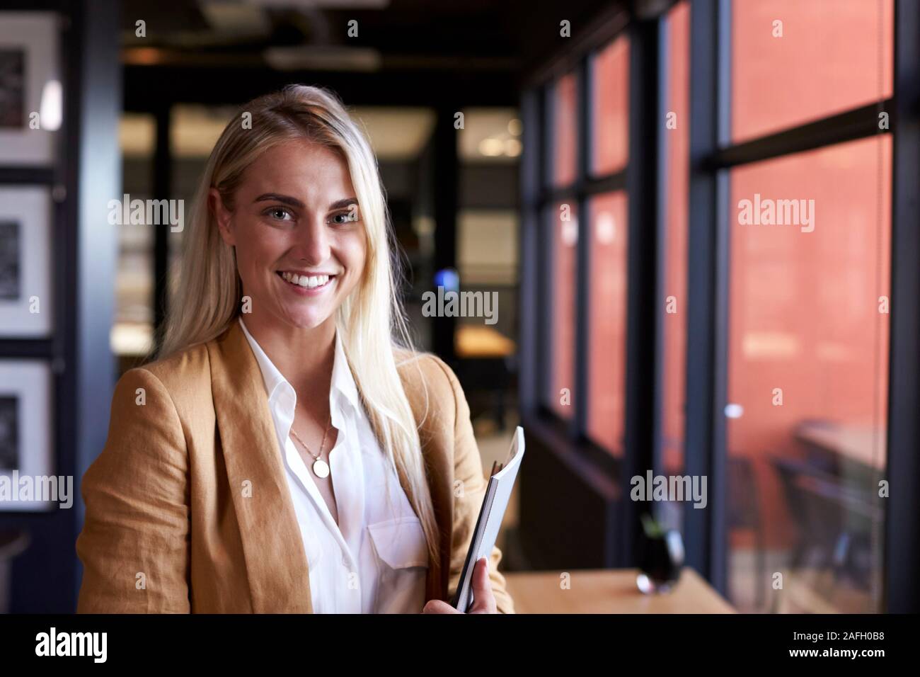Bianco millenario bionda imprenditrice sorridere alla telecamera tramite la finestra in un ufficio, close up Foto Stock