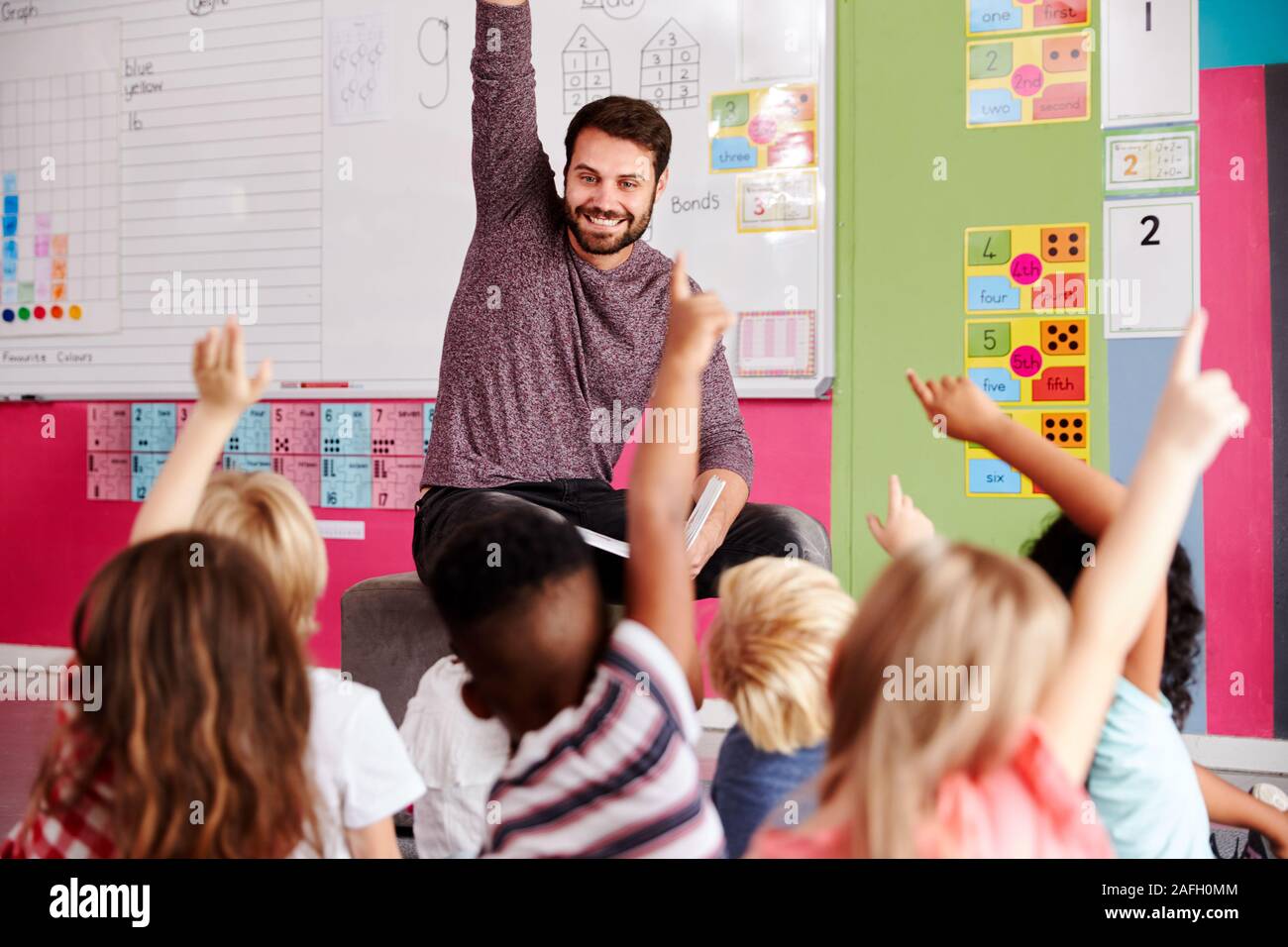 Gli alunni elementari alzando le mani per rispondere alla domanda come maschio insegnante legge la storia in aula Foto Stock