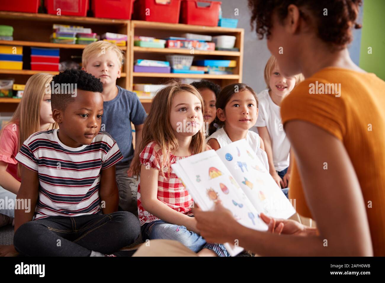 Gruppo di scuola elementare alunni seduti sul pavimento ascoltando insegnante femminile Leggi la storia Foto Stock