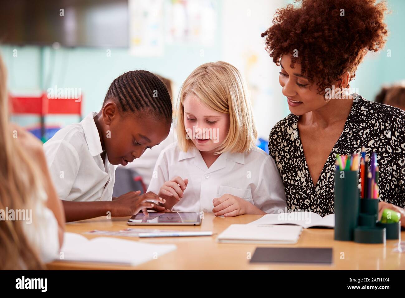 Insegnante femmina con due scuole elementari gli alunni indossano uniformi utilizzando tavoletta digitale alla scrivania Foto Stock