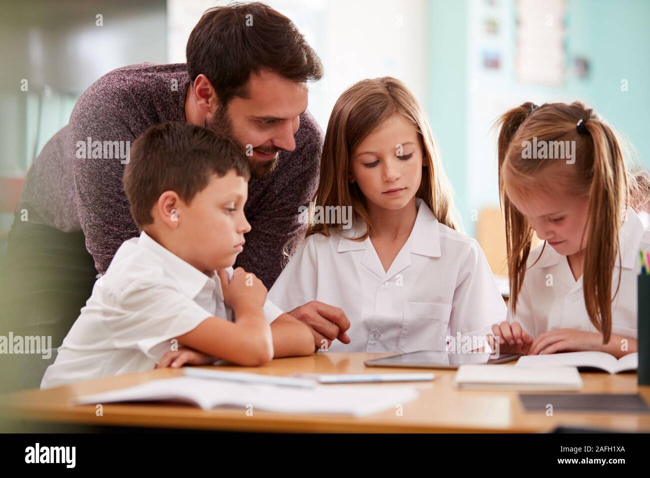 Insegnante di sesso maschile con tre scuole elementari gli alunni indossano uniformi utilizzando tavoletta digitale alla scrivania Foto Stock