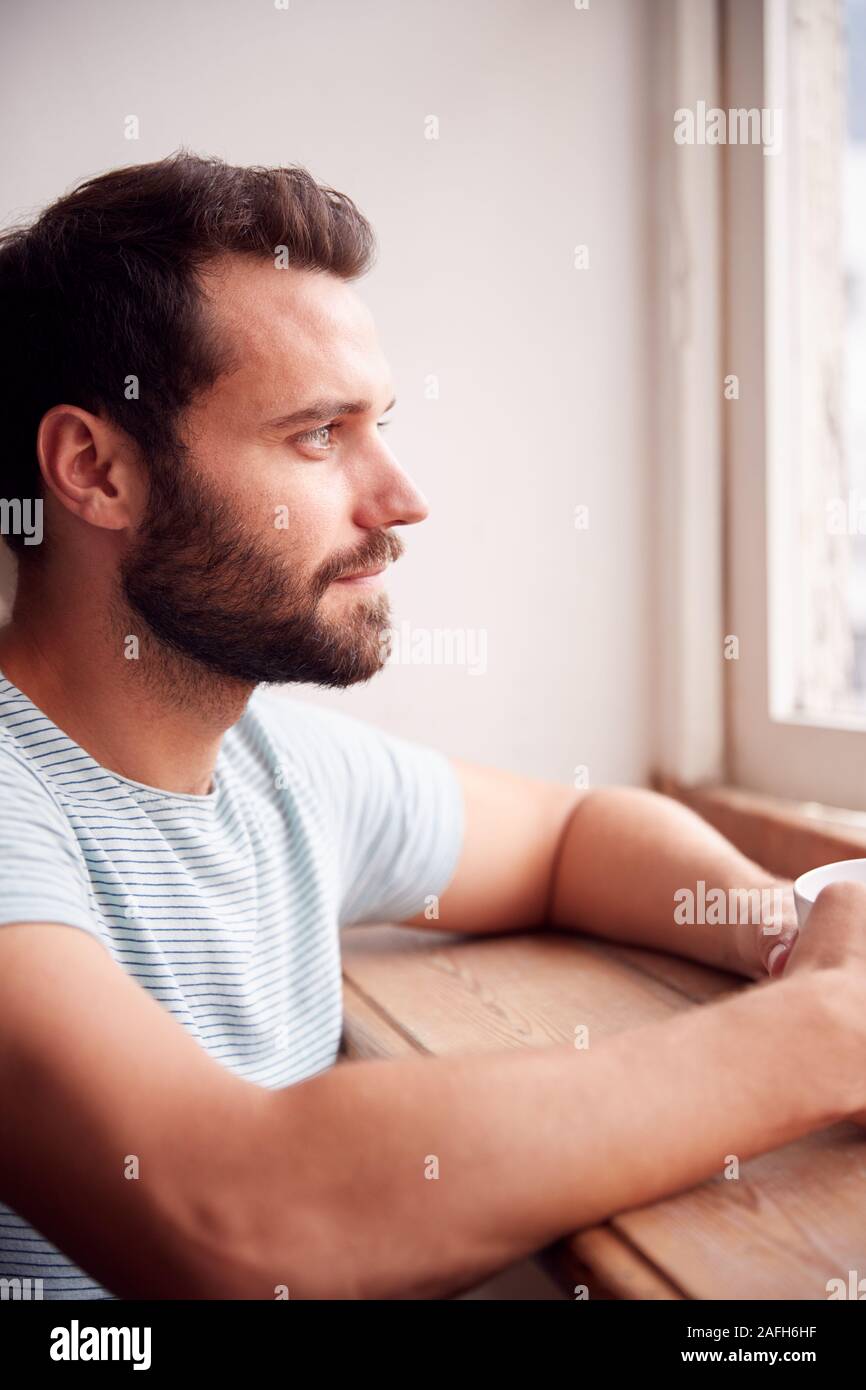 Giovane uomo rilassante in appartamento loft guardando fuori della finestra con bevanda calda Foto Stock
